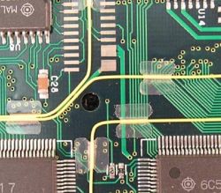 Zbliżenie na płytkę PCB z żółtym przewodem kynarowym przyklejonym za pomocą przezroczystych naklejek.