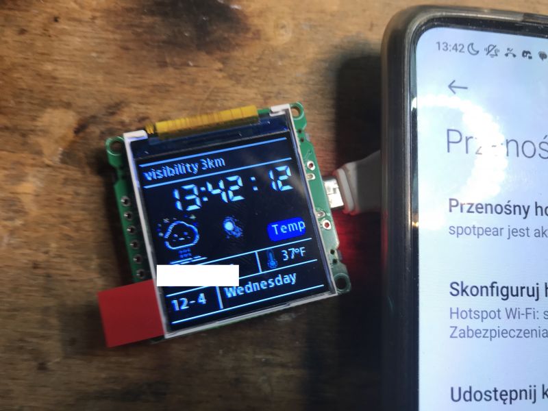 ESP32-C3 Display Module Next to Smartphone Miniature ESP32-C3 display module showing weather information next to a phone.