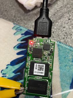 Close-up of an open SSD circuit board connected via USB cable, with visible chips and a red LED indicator.