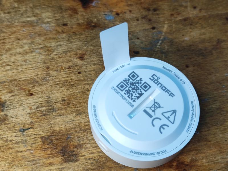 Tył urządzenia Sonoff SNZB-01P na drewnianym tle z widoczną baterią.