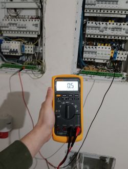 Osoba trzymająca miernik napięcia przed otwartą rozdzielnicą elektryczną.