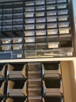 Szafka z plastikowymi szufladkami na drobne elementy elektroniczne, częściowo opisana.