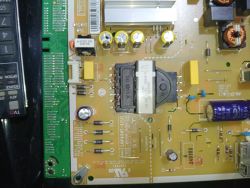 Zbliżenie na płytę zasilacza telewizora z kondensatorami i transformatorem, obok fragment zielonej PCB
