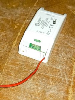 A white Connex Connect Smart WiFi Switch 10A module with a red wire attached, resting on a wooden surface.