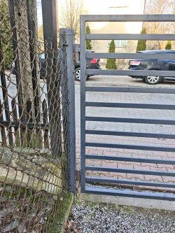 Zbliżenie na zawias i słupek metalowej furtki przy ogrodzeniu z siatki, z wąskim prześwitem.