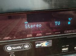 Wyświetlacz amplitunera z ustawieniem Stereo oraz TV, z widocznymi ikonami Spotify i dts.