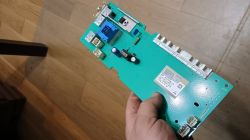 Osoba trzyma w ręce płytkę elektroniki modułu pralki nad drewnianą podłogą.
