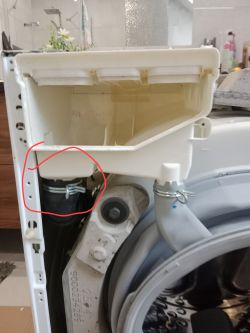 Zaznaczona metalowa obejma na gumowym wężu pod dozownikiem pralki.
