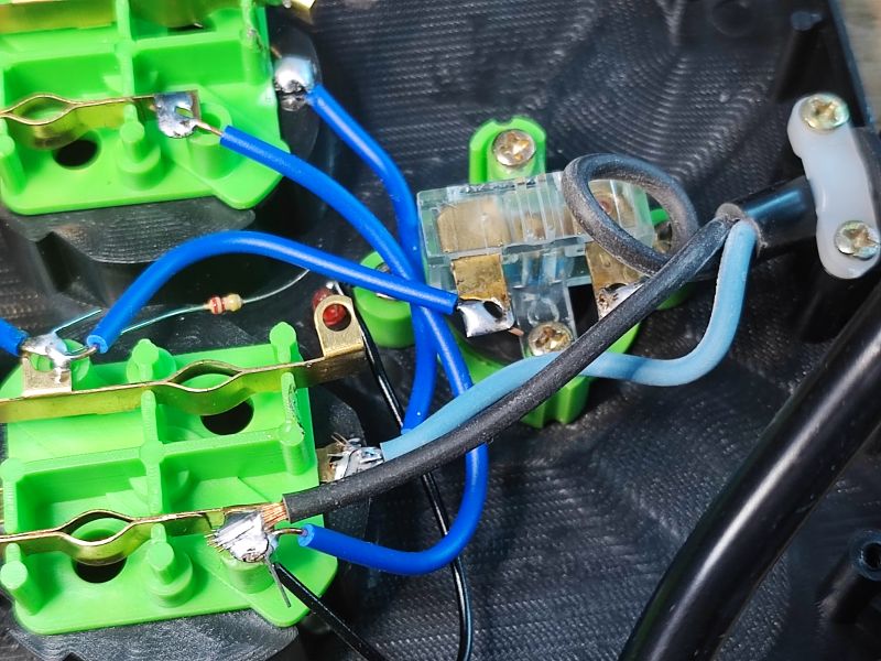 Wnętrze listwy zasilającej z widocznym chaotycznym prowadzeniem przewodów, lutowaniami i elementami mocującymi na zielonych plastikowych podstawach.
