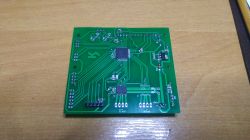 Zielona płytka PCB z zamontowanymi elementami elektronicznymi, w tym układami scalonymi i kilkoma rezystorami, leży na drewnianym blacie.
