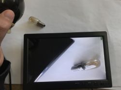 Small black monitor shows a close-up of a light bulb; another bulb and a hand are visible on the desk.