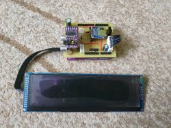 A custom radio module with ESP32 and an LCD display lying on a carpet, connected by a cable.