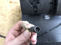 Close-up of a mini-DIN plug held in a hand, with a black device housing in the background.