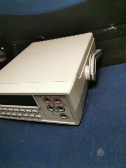 Biały miernik elektroniczny z wyświetlaczem i złączami, umieszczony na niebieskiej powierzchni.