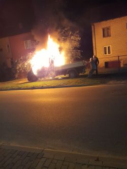 Płonący samochód dostawczy nocą na trawniku obok domów, dwóch mężczyzn obok pojazdu