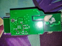 Green PCB with visible traces, pads, pin labels, and a drop of white adhesive or resin in the top right corner.