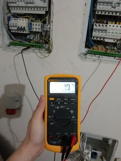 Multimetr wskazujący napięcie 1.9 mV z rozdzielnicą elektryczną w tle.
