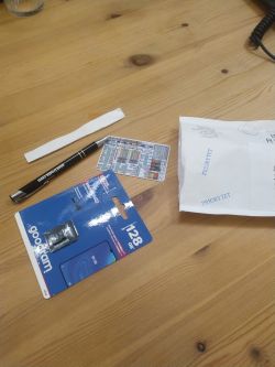 On a wooden table, there is a pen, a 128 GB memory card in packaging, a resistor color code chart, and an envelope marked PRIORYTET.