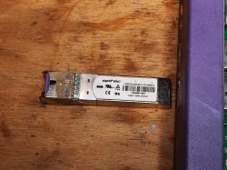 Moduł SFP optyczny marki Optic leżący na drewnianym blacie, z widocznymi śladami korozji.