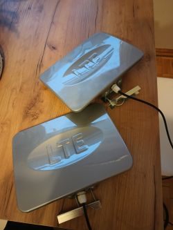 Dwie szare anteny LTE leżące na drewnianym stole z widocznymi uchwytami montażowymi i kablami.