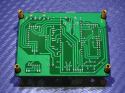 Bottom view of a green PCB with traces and through-hole pads, four gold standoffs in the corners