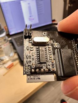Close-up of a black electronic circuit board held in hand with components, including a quartz crystal and pins.