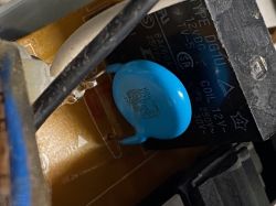 Zbliżenie wnętrza urządzenia: płytka PCB, niebieski dyskowy element i czarny kondensator z nadrukiem 12V