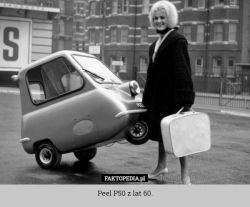 Kobieta stoi obok małego samochodu Peel P50 z lat 60., trzymając walizkę.