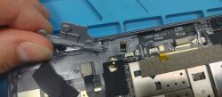 Wnętrze tabletu z widoczną płytą i taśmami; dłoń trzyma szary element plastikowy przy krawędzi