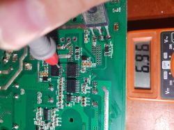 A hand holding a multimeter probe touches a point on a circuit board, with the multimeter display showing a reading of 6.96.