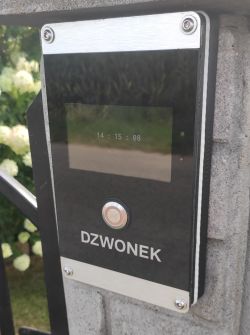 Panel dzwonka z ekranem dotykowym za szkłem, zamontowany na murku ogrodzenia