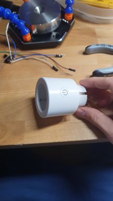A hand holding a white electronic plug with a power button on a workshop table.