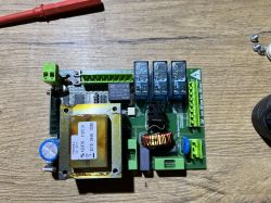 Zbliżenie na płytkę drukowaną z elektronicznymi komponentami, takimi jak przekaźniki i transformator.