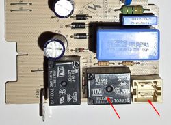 Zbliżenie na płytkę drukowaną z elementami elektronicznymi, w tym przekaźnikami, kondensatorami i złączem. Dwa czerwone wskaźniki wskazują na przekaźnik i złącze.