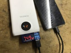 Powerbank Romoss ładuje smartfona, między urządzeniami podłączony miernik USB pokazujący napięcie 7,49 V.