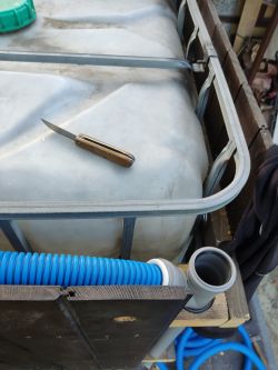 Knife on a white plastic tank next to a blue hose and gray PVC pipe.