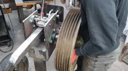 Close-up of a metal kinetic log splitter with a toothed rack and a large grooved pulley wheel in a workshop