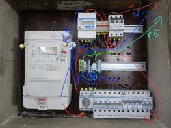 Zdjęcie instalacji elektrycznej z widocznym licznikiem i rozdzielnicą z podpisanymi przewodami.