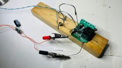 Simple electronic setup on a wooden board with a relay, capacitor, and wires with clips.