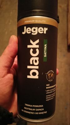 Ręka trzymająca puszkę czarnego sprayu Jeger Satyna, przeznaczonego do malowania powierzchni.