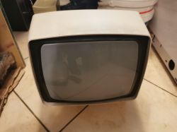 Stary telewizor kineskopowy o białej obudowie, stojący na podłodze na tle innych sprzętów.