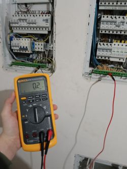 Ręka trzymająca multimer z wyświetlaną wartością rezystancji 0,2 Ω przed otwartą skrzynką elektryczną.