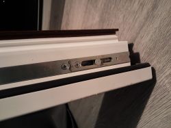 Zbliżenie na metalowy mechanizm zasuwnicy okiennej zamontowany w białej ramie okna