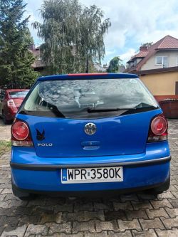 Blue Volkswagen Polo car seen from the back, parked on a driveway surrounded by houses and trees.