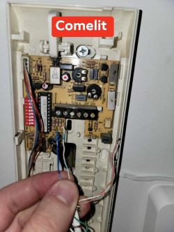 Close-up of the inside of a Comelit intercom showing a circuit board and disconnected wires held by a hand.