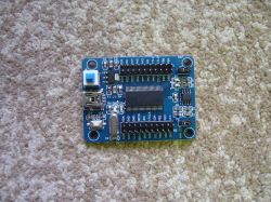 Moduł elektroniczny z niebieskim przyciskiem, złączem USB i różnymi komponentami na niebieskiej płytce PCB.