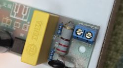 Zbliżenie płytki PCB z żółtym kondensatorem, dwoma rezystorami i niebieską złączką śrubową