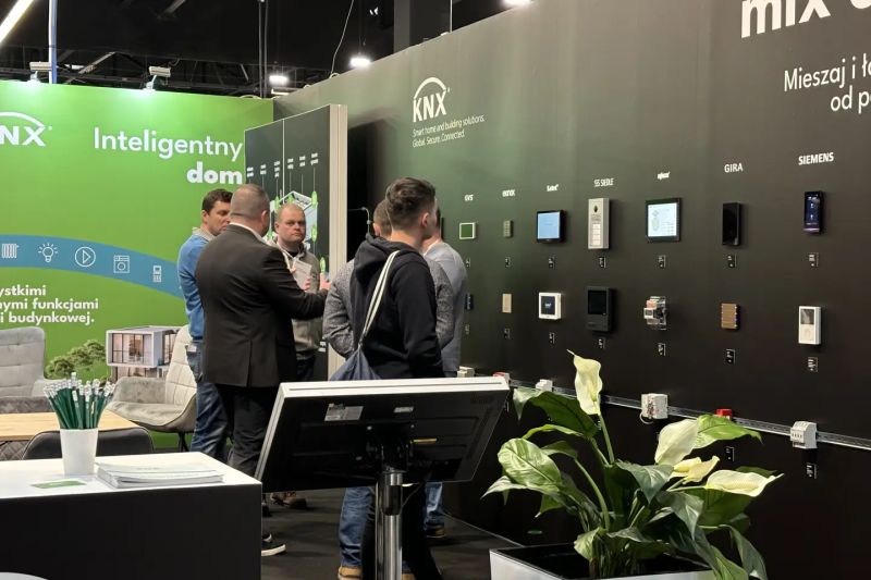 KNX trade fair booth; people view a wall display of smart home control panels and devices.