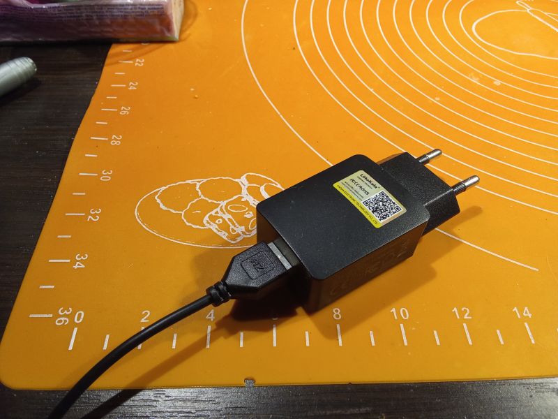 Czarna ładowarka USB z podłączonym kablem na pomarańczowej macie z oznaczeniami miarowymi.