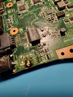 Zbliżenie na płytę główną laptopa Asus X53S z widocznymi elementami elektronicznymi i uszkodzeniem w pobliżu gniazda ładowania.